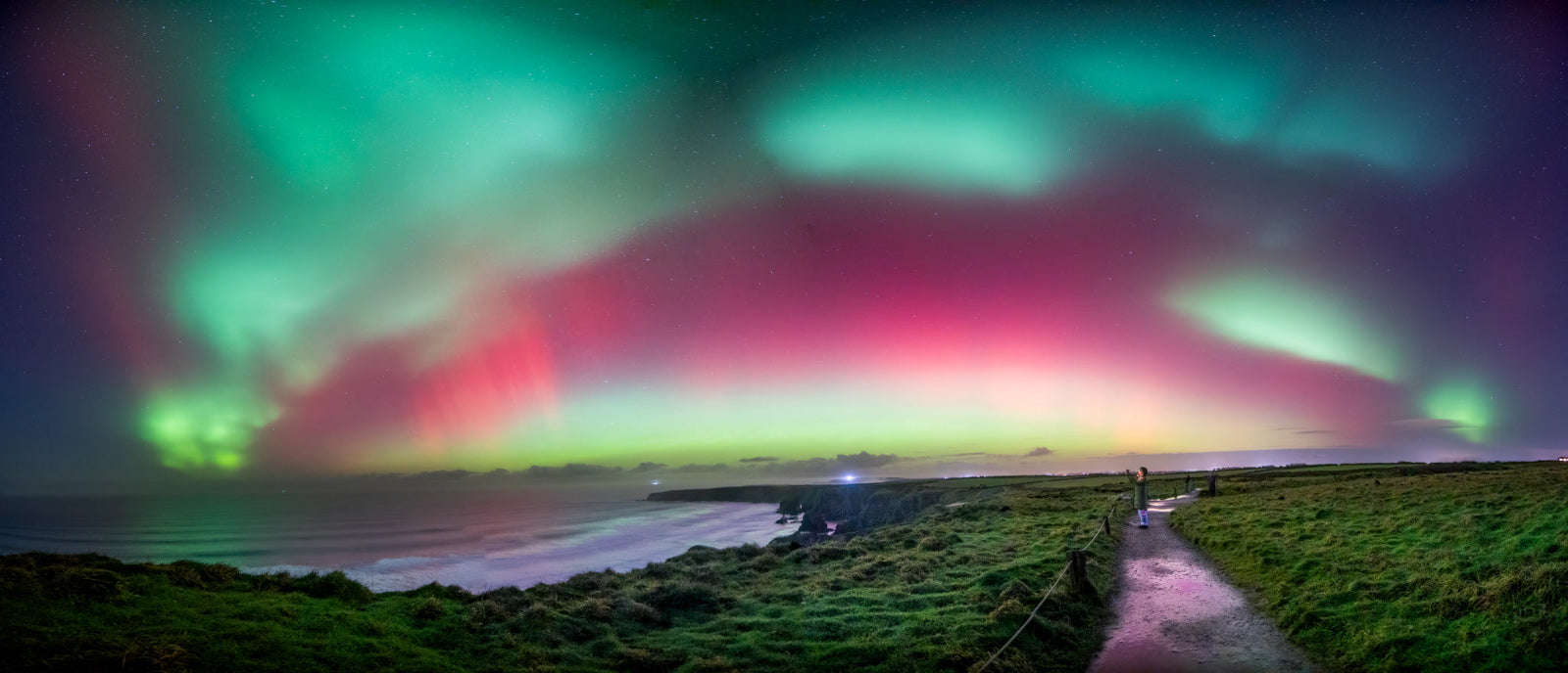 Aurora borealis over a coastal landscape with a person walking along a path.