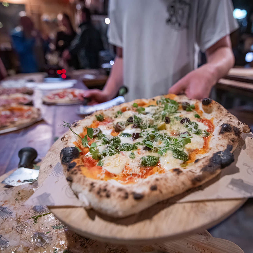 Person holding a pizza with various toppings on a wooden board in a casual setting.